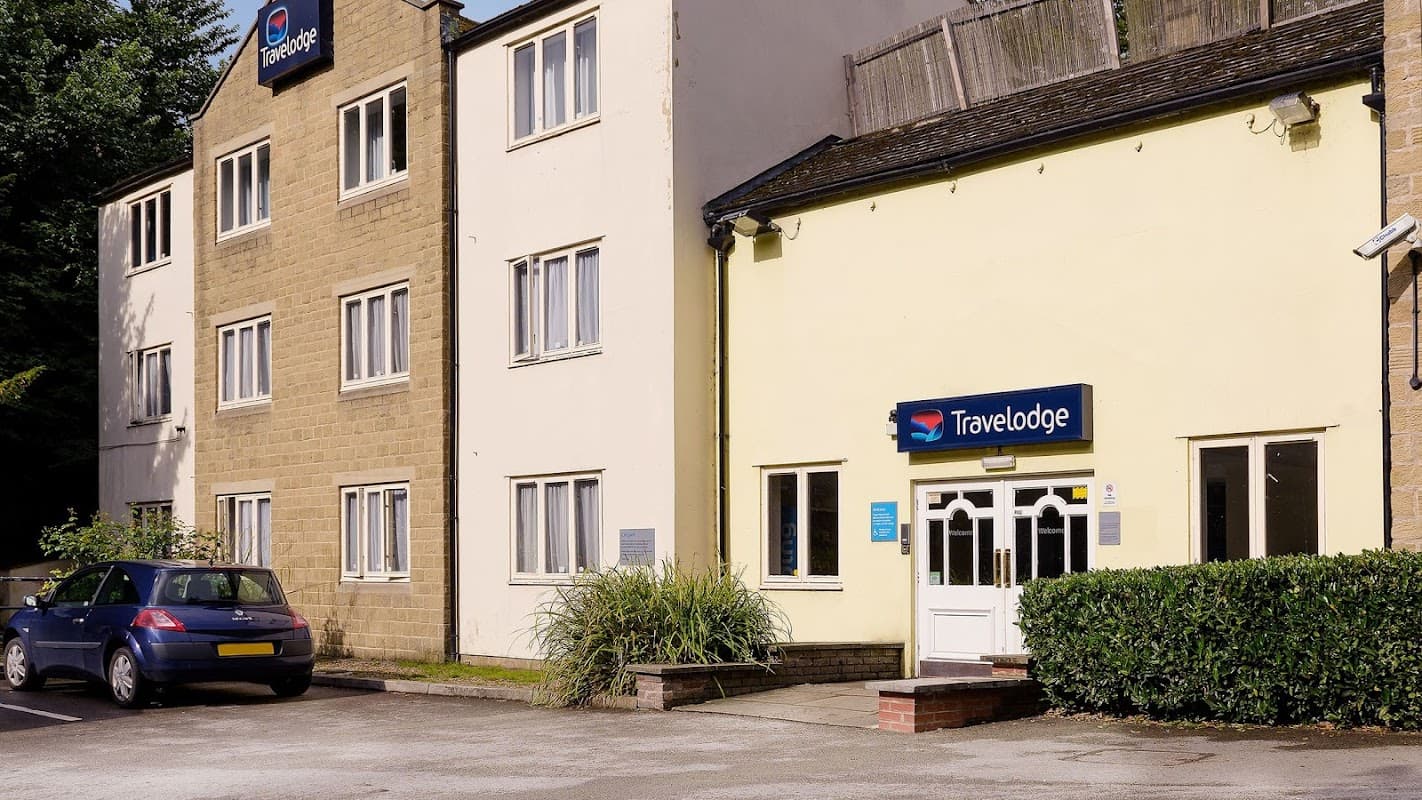 Travelodge building with a yellow exterior, blue signage, and parked car in front, surrounded by greenery.