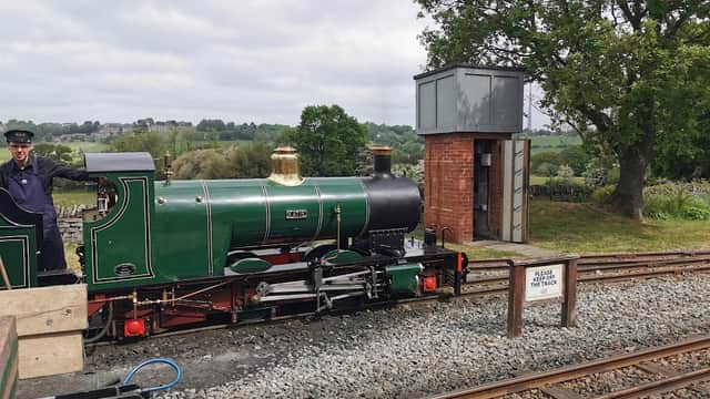 A green steam locomotive at a railway station with a conductor, surrounded by tracks and greenery.