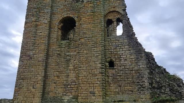 Ruined stone tower with arched openings, set against a cloudy sky, surrounded by grassy terrain.