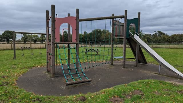 Playground equipment with a climbing net, slide, and swings on grassy field under a cloudy sky in West Haddlesey.
