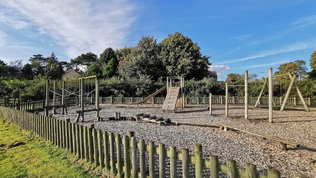 Play park with climbing frames, slides, and a gravel surface, surrounded by wooden fencing and greenery under a blue sky.