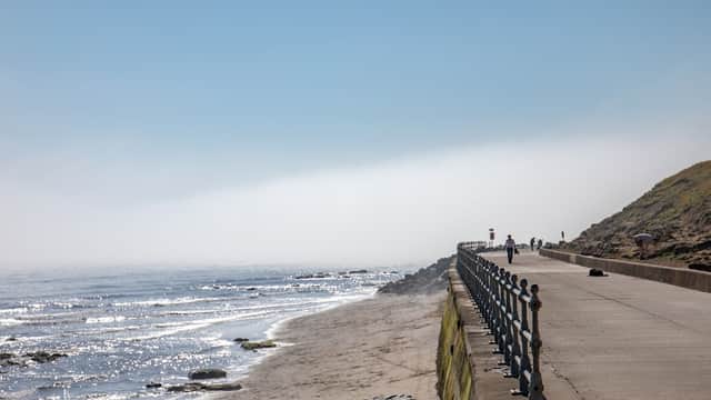 Coastal path alongside a sandy beach with gentle waves, under a clear blue sky and distant fog.