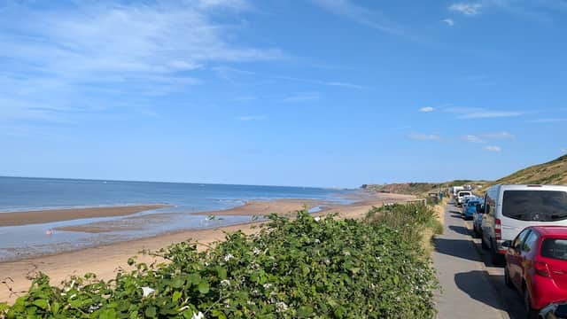 Car park lined with vehicles beside a sandy beach and calm sea under a clear blue sky in Whitby, Yorkshire.