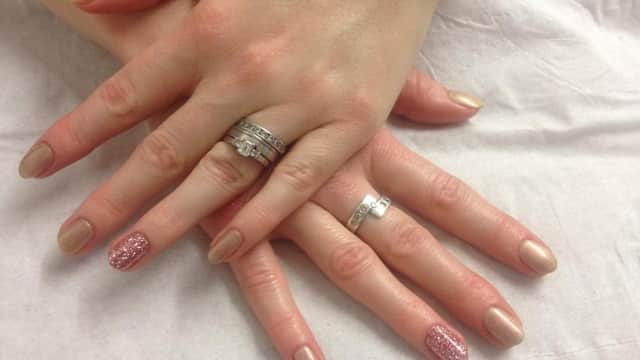 Two hands with manicured nails, one featuring glitter, adorned with multiple rings, resting on a white surface.