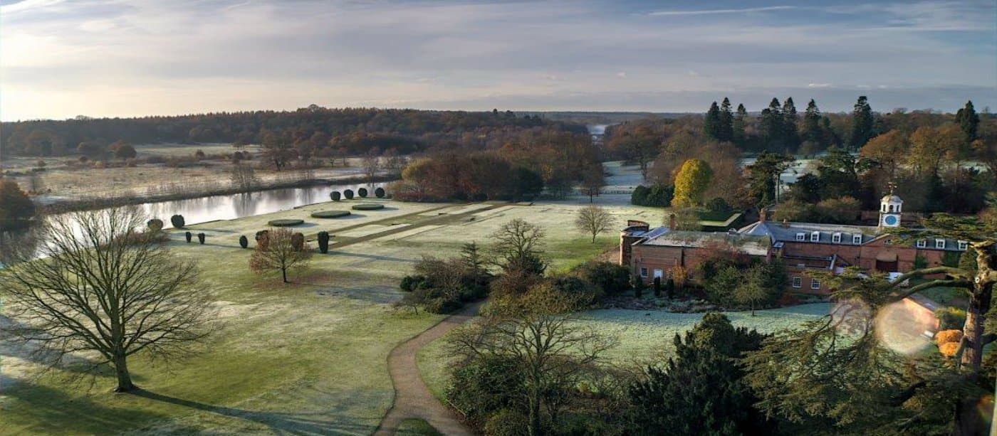 Expansive parkland with frosted grass, a winding path, a lake, and a historic brick building surrounded by trees.