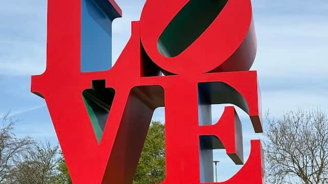 Large red and blue "LOVE" sculpture in a grassy area with trees and a clear blue sky in the background.