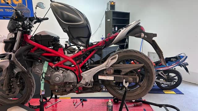 Motorcycles on a repair stand in a bike shop, tools and equipment visible in a well-lit workshop setting.