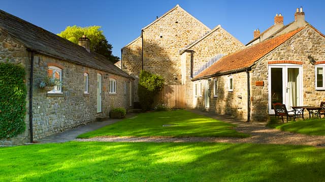 Stone buildings with slate roofs, a green lawn, and a small seating area under clear blue skies in Crakehall, Yorkshire.