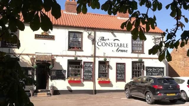 Traditional pub with a white exterior, red roof, and black signage, surrounded by greenery and parked cars.