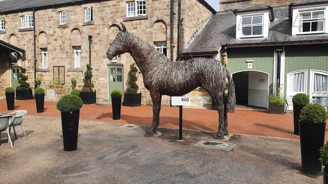A large straw horse sculpture stands in a courtyard surrounded by potted plants and a brick building.