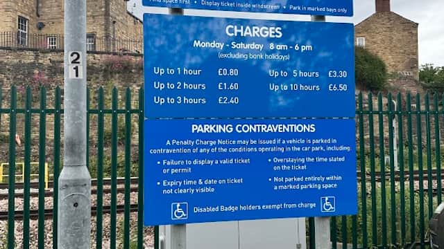 Sign for Wellington Road Car Park in Dewsbury, listing charges and parking regulations, with a cloudy sky above.