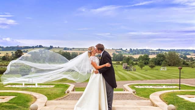 A bride and groom kiss on a stone pathway, with lush green fields and blue skies in the background.
