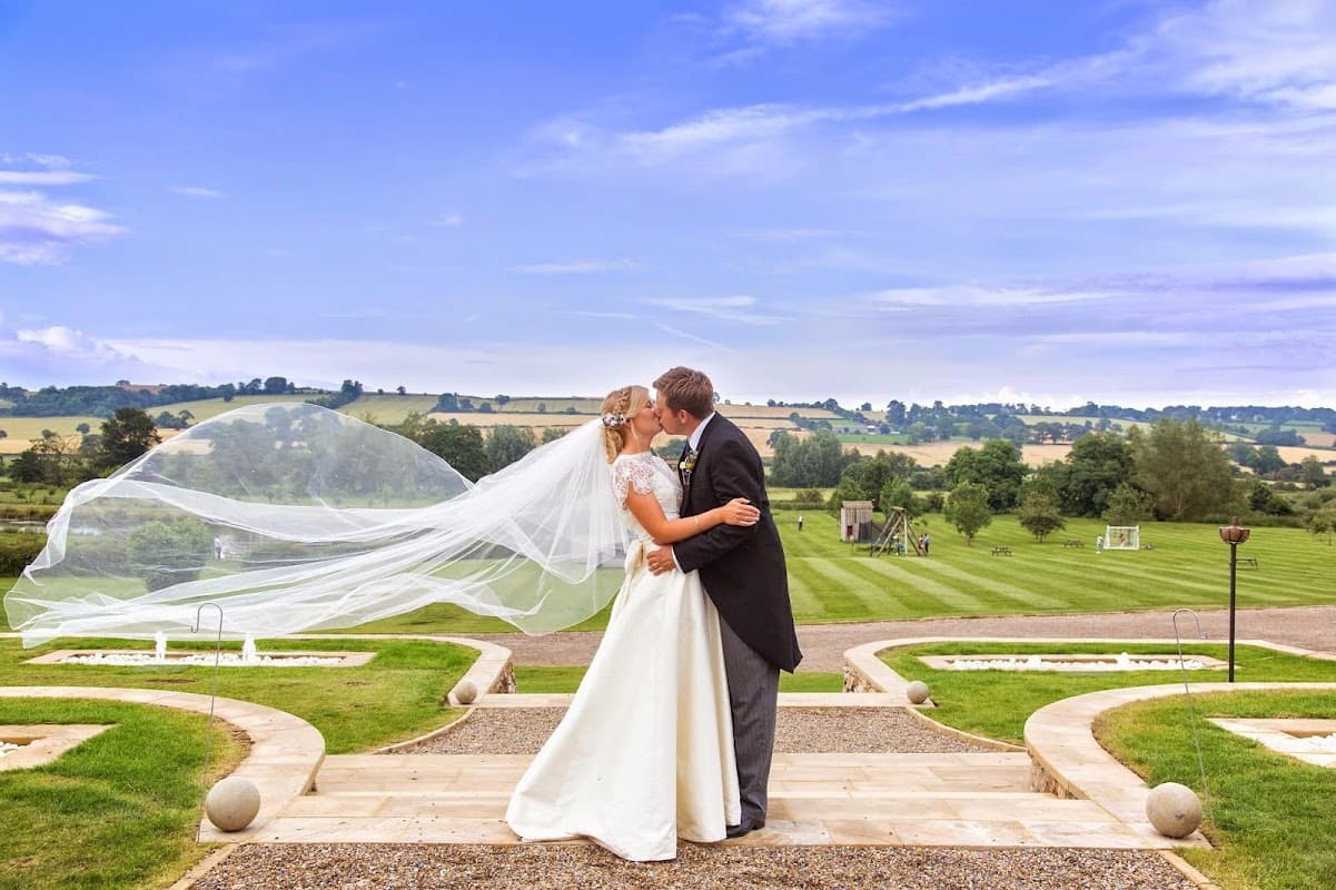 A bride and groom kiss on a stone pathway, with lush green fields and blue skies in the background.