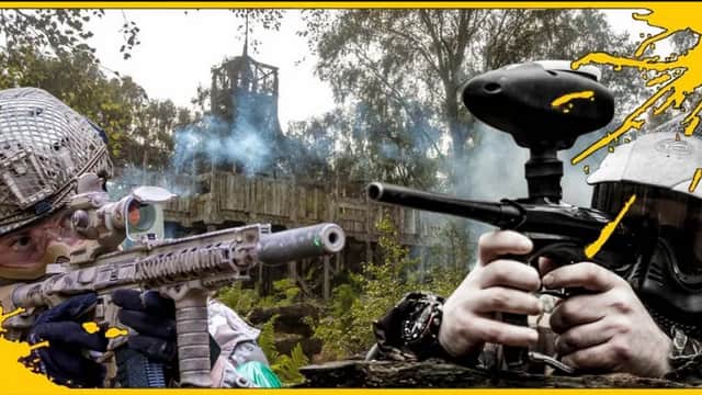 Two players in tactical gear aiming paintball guns at each other, with a wooden structure and smoke in the background.