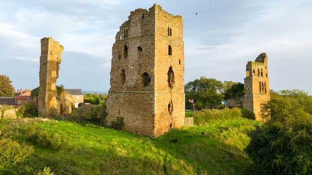 Ruins of Sheriff Hutton Castle with grassy mounds and trees, under a blue sky with scattered clouds.