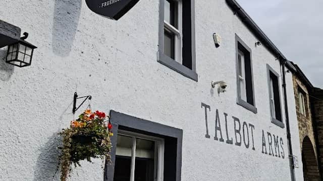 White-washed exterior of Talbot Arms pub, featuring a dog sign, window boxes with flowers, and a cobblestone entrance.