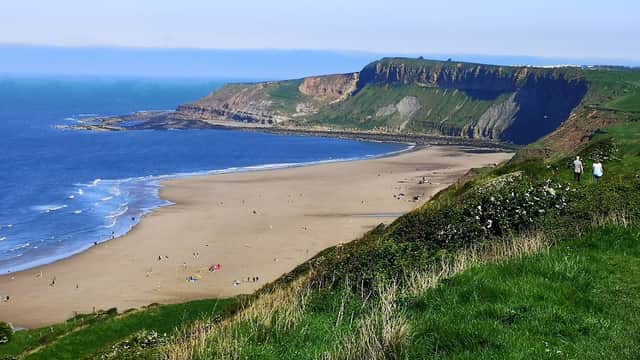 Cayton Bay with sandy beach, cliffs, and green hills under a clear blue sky, featuring people enjoying the shore.