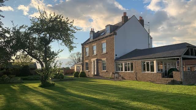 Luxury cottage with a spacious lawn, trees, and a two-story brick exterior under a blue sky with fluffy clouds.