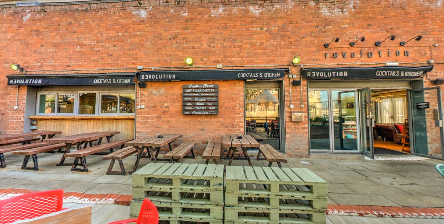 Outdoor seating area with wooden benches, a bar entrance, and a brick wall featuring the "Revolution" sign.