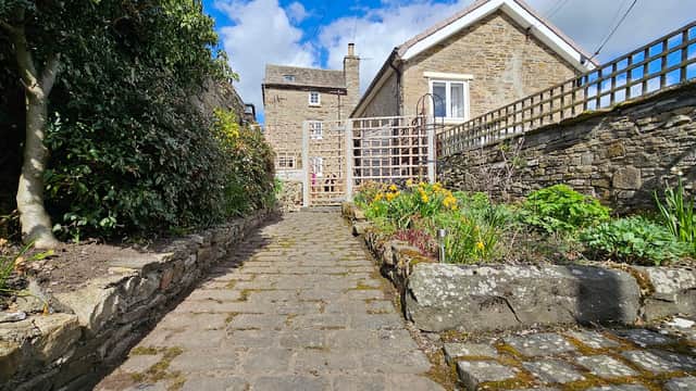 Pathway leading to Puzzle Cottage, flanked by stone walls and vibrant garden flowers under a blue sky.