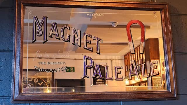 Framed sign featuring "Magnet Pale Ale" with a red magnet illustration, set against a rustic pub interior.