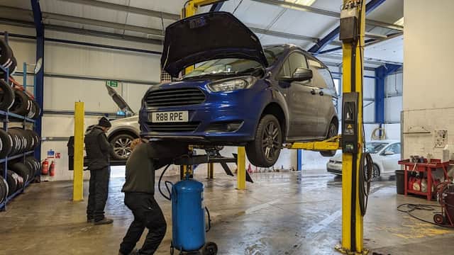 A blue car on a lift in a garage, with a mechanic working underneath and tyres stacked in the background.