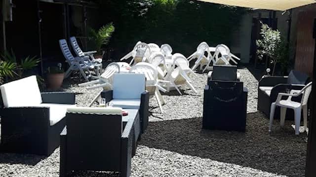 Outdoor seating area with lounge chairs and tables on gravel, surrounded by greenery and shaded by a canopy.