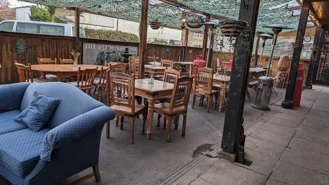 Outdoor seating area with wooden tables and chairs, a blue sofa, and a canopy overhead, in a rustic bar setting.