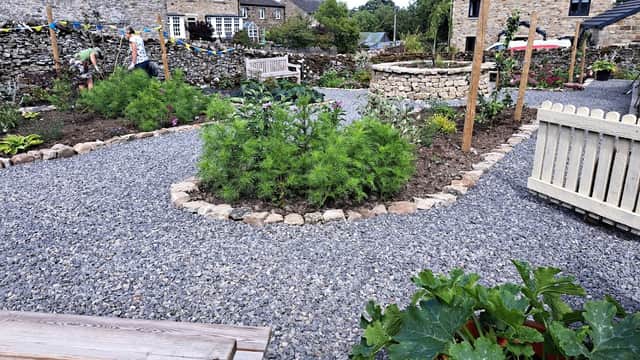 Lush community garden with gravel paths, stone walls, benches, and various plants in Bellerby, Yorkshire.