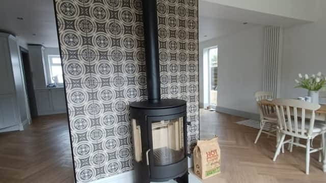 Modern wood stove with glass door, set against a patterned wall, in a spacious, light-filled room with wooden flooring.