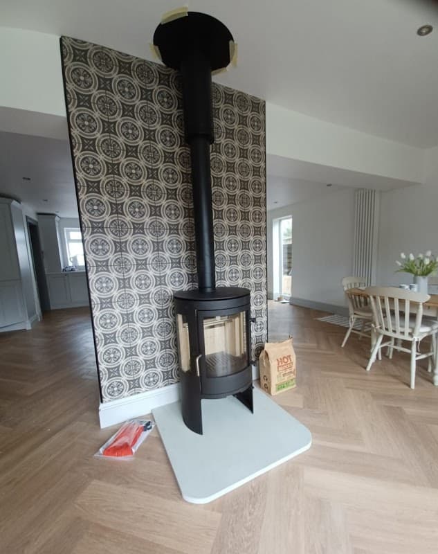 Modern wood stove with glass door, set against a patterned wall, in a spacious, light-filled room with wooden flooring.