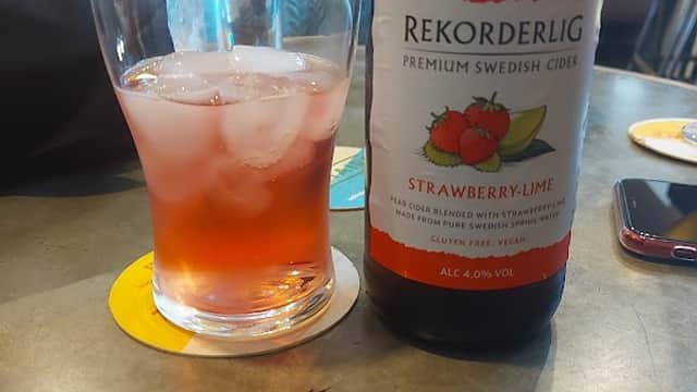 A glass of ice with a pink drink beside a bottle of Rekorderlig Strawberry-Lime cider on a table.
