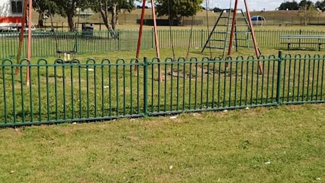 Swings in a grassy area surrounded by a green fence, with trees and blue sky in the background.