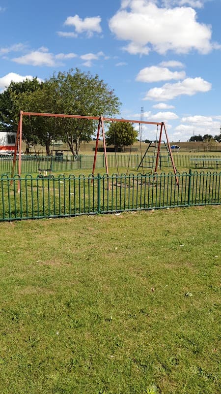 Swings in a grassy area surrounded by a green fence, with trees and blue sky in the background.