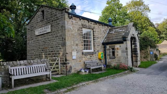 Stone building with a sign reading "Village Hall for Hire," surrounded by trees and benches, in Leathley, Yorkshire.