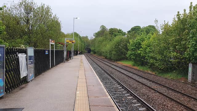 Car park beside railway tracks, trees lining the area, and platform with signage under overcast sky.