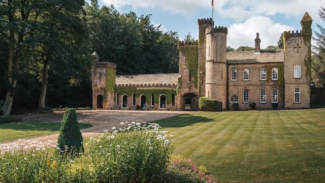 Victorian-style castle surrounded by lush greenery and manicured lawns, with a flag flying and flower beds in the foreground.