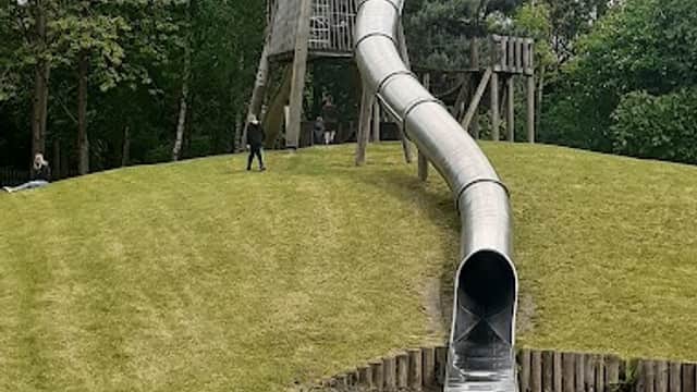 Large silver slide winding down a grassy hill, surrounded by trees and a playground area in Hemsworth Waterpark.