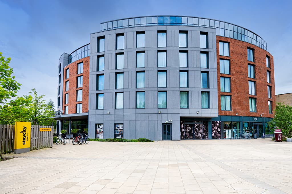 Modern hotel building with a mix of grey and red brick, large windows, and a spacious plaza with bicycles parked outside.
