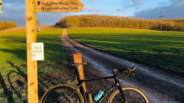 A bicycle leans against a wooden signpost on a dirt path surrounded by green fields and trees under a blue sky.