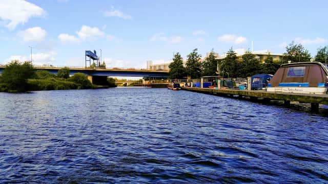 View of a river with boats and a car park in the background, under a clear blue sky with scattered clouds.