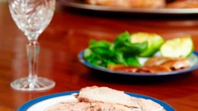 Sliced meat on a plate with a crystal glass and garnishes, with a whole roasted pig in the background.