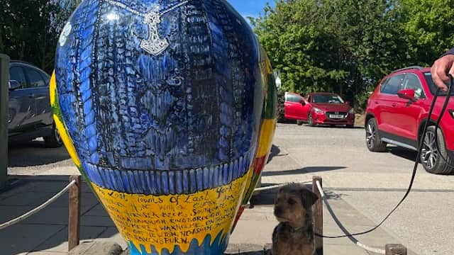 Colorful bird sculpture with intricate designs, a small dog sitting nearby, and parked cars in the background.