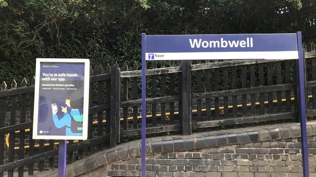 Wombwell bus stop sign with a travel app advertisement nearby, surrounded by greenery and a stone wall.
