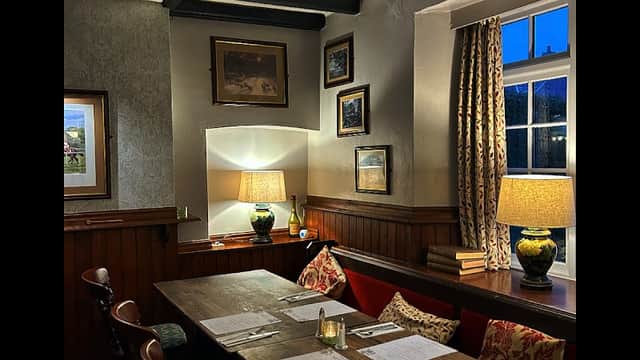Cozy bar interior with wooden beams, a long table set for dining, warm lighting, and decorative wall art.
