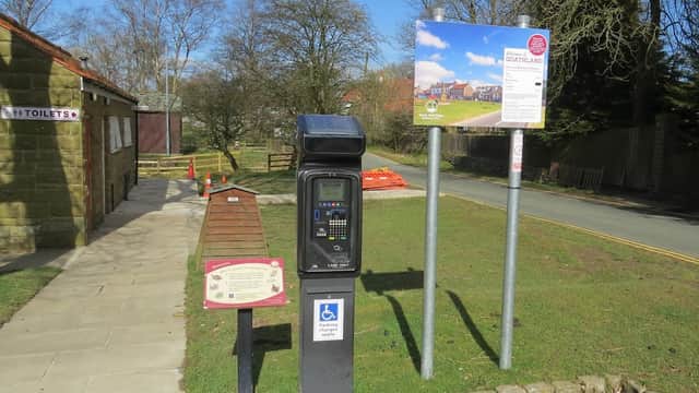 Pay & Display parking machine and information sign near a path, with trees and a road in the background.