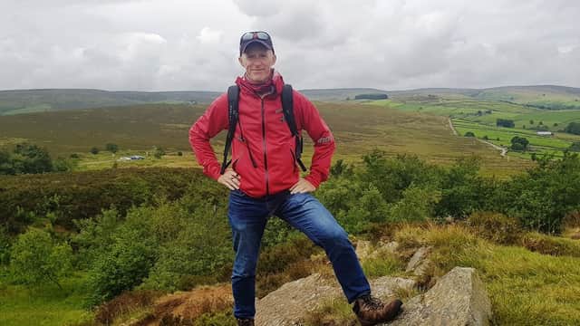 A man in a red jacket stands confidently on a rock, surrounded by lush green hills and a cloudy sky in Yorkshire.