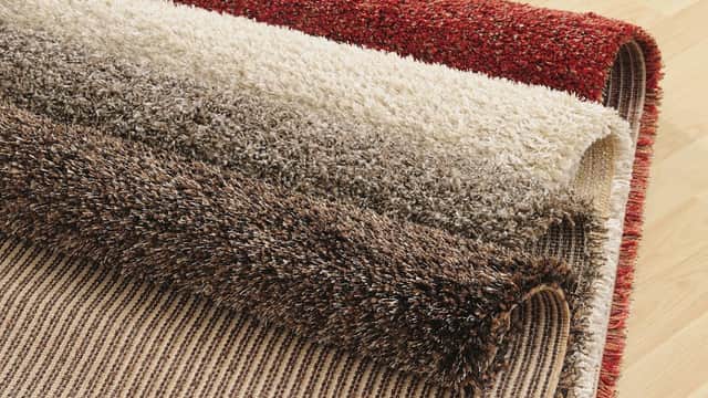 Four rolls of plush carpets in varying shades of red, cream, brown, and beige, displayed on a wooden floor.