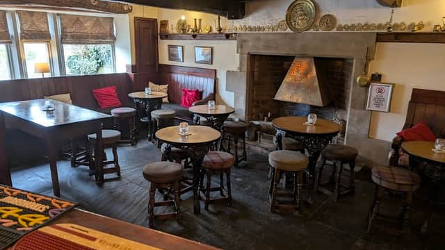 Cozy pub interior featuring wooden beams, tables, stools, and a stone fireplace with decorative elements.