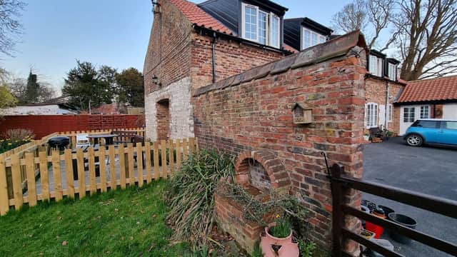 Brick wall with an arch, green grass, and holiday cottages with windows, surrounded by a wooden fence and trees.